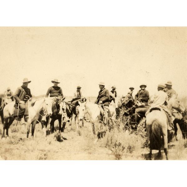 A photo of ten men on horseback. They are wearing hats. There are short bushes in the foreground.