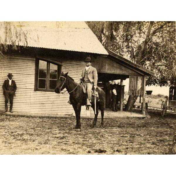 Une photo d'un homme en costume sur un cheval foncé. Il tient un fouet à la main. Il y a un bâtiment avec un revêtement et un porche couvert derrière lui. Un autre homme s'appuie contre le mur du bâtiment. 