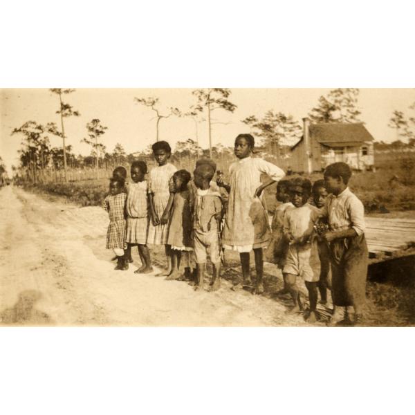 A photo of a group of children standing beside a dirt road. There is a building behind them and trees in the background.