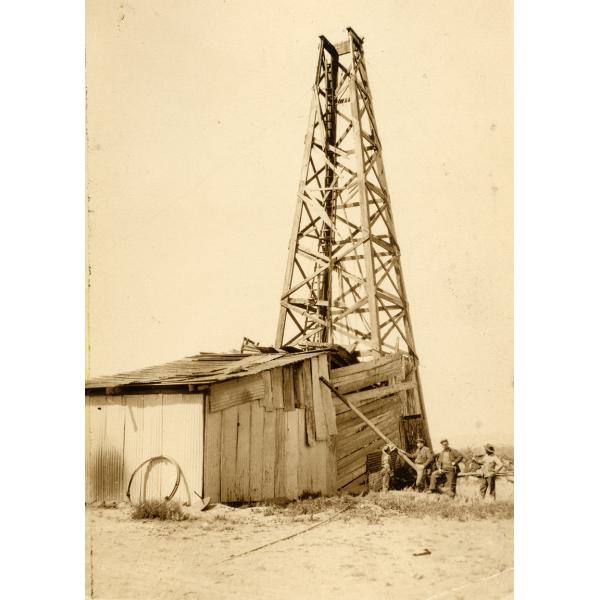 Four men standing in a dusty field beside a wooden oil rig with four sides. The bottom of the rig, with the machinery inside it, is covered in wooden boards and metal sheeting.