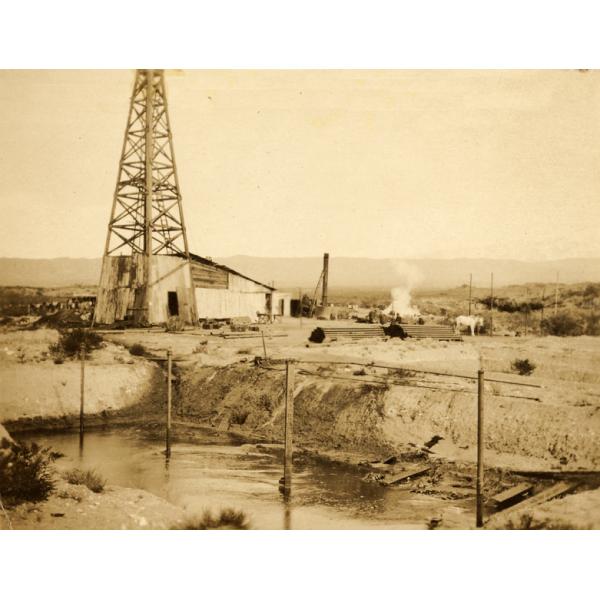 A photo of an oil rig behind a large dug hole with water in it and vertical posts. There is a pile of casing beside the rig with two men and a white horse standing beside it. There are hills in the background.