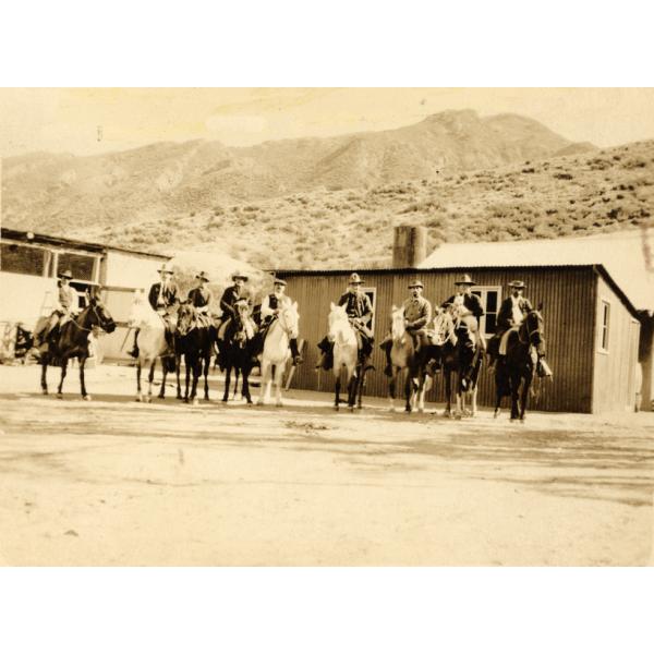 A photo of nine International Drillers sitting on horses and wearing hats. There are two single-story buildings behind them and rolling hills in the background.