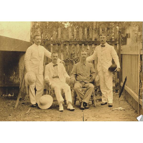 Two men sitting in chairs with a man standing on either side. They are wearing white and light coloured clothing. Each man is holding a hat or resting one on the ground. They are standing in front of a fence with vines on it. 