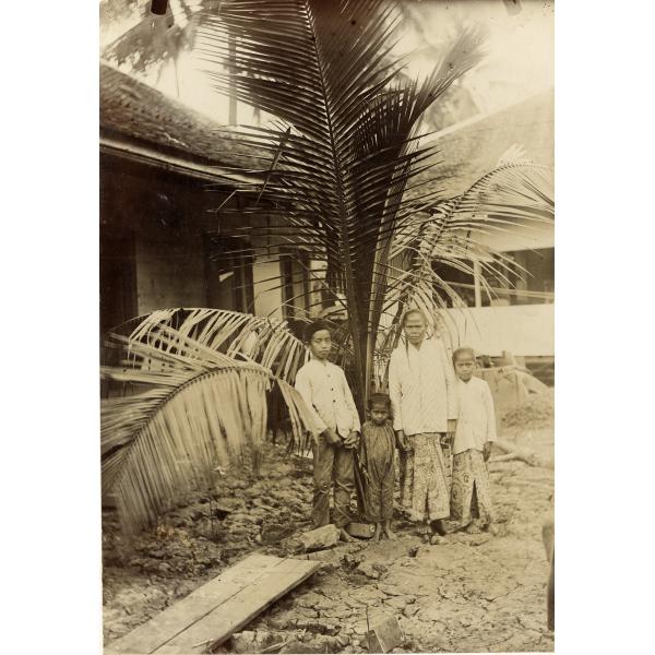 A woman and three children standing in front of palm fronds and a building on cracked dirt. The woman and two oldest children are wearing white shirts, while the youngest is wearing a dark outfit. 