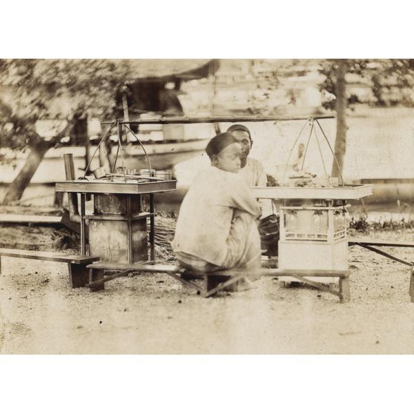 Two men sit on opposite sides of a pair of food stands. There is cookware on top of both stands, and there is a wooden bar attached to wire hangers that run down to the stands.