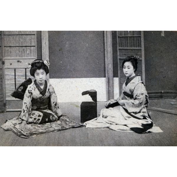 Une photo de deux fillettes vêtues de vêtements japonais, assises sur des nattes pliées. Il y a une boîte foncée qui est ouverte et posée entre elles. Il y a un mur avec des nattes derrière elles.