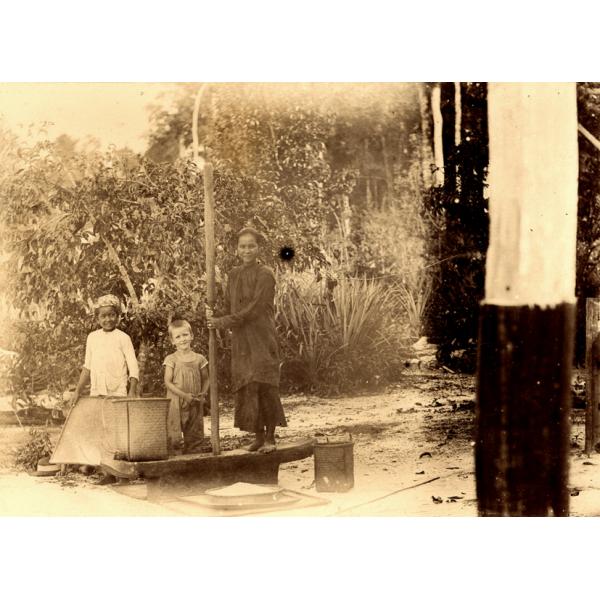 A photo of a woman standing on a platform with two children. She is holding a long pole and there is a basket on either side of her. There are bushes in the background. 