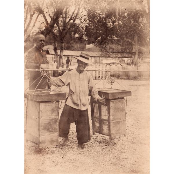 A man balancing a pole across his shoulders, with a wooden table on either side. A man is standing behind him. There is a picket fence and a couple of trees in the background. 