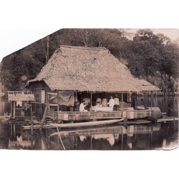 A photo of a bungalow with a thatched roof, floating on a raft. There is a group of people on the deck and trees in the background.  