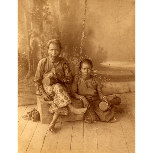 A photo of an old woman sitting on a wooden box with her legs crossed. A younger woman sits on the floor made of wooden boards beside her. There is a screen behind them with trees on it.  