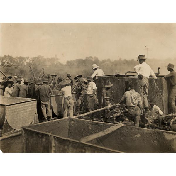 Bloss Sutherland and George York look on as a group of native workers stir cement in a metal barrel with sticks. There is another barrel beside them.  