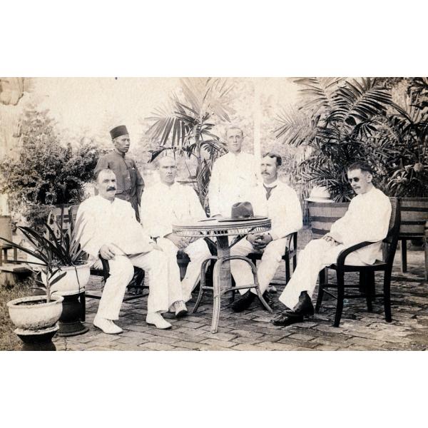 A photo of five International Drillers, wearing white, around a table with a hat on it. A servant stands behind them. There are trees in the background. 