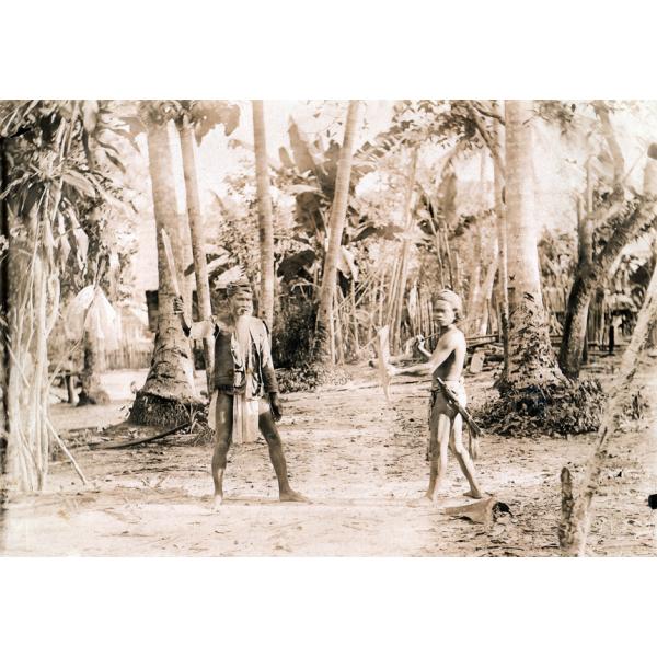 A photo of a man and a boy dressed in the traditional clothing of the Batak tribe. The man is holding a blade and the boy may be as well. They are standing in front of a number of trees.  