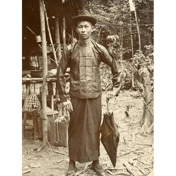 Photo d'un Chinois portant un chapeau, une chemise boutonnée et une jupe. Il tient un parapluie fermé à ses côtés. Il y a une structure avec des poteaux en bois et un toit derrière lui. 