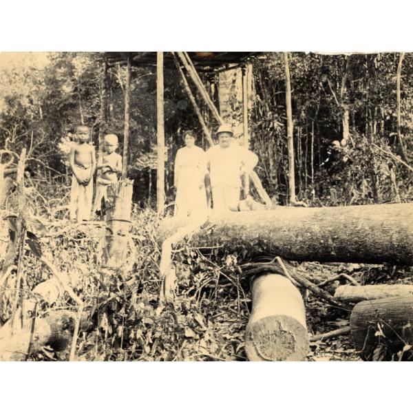 William McRae standing in the jungle with a woman and two native children. Felled trees occupy the foreground and a roughly built structure of timbers appears in the back.