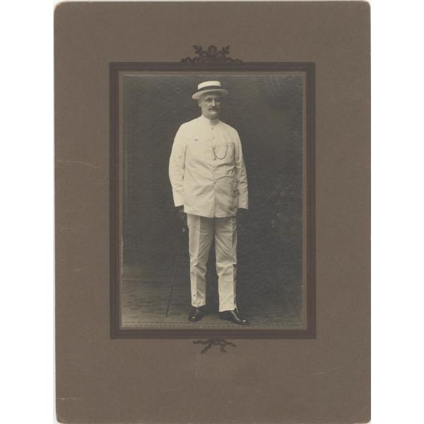 William McRae standing in a white suit and panama hat. Photograph is placed in a brown matte. 