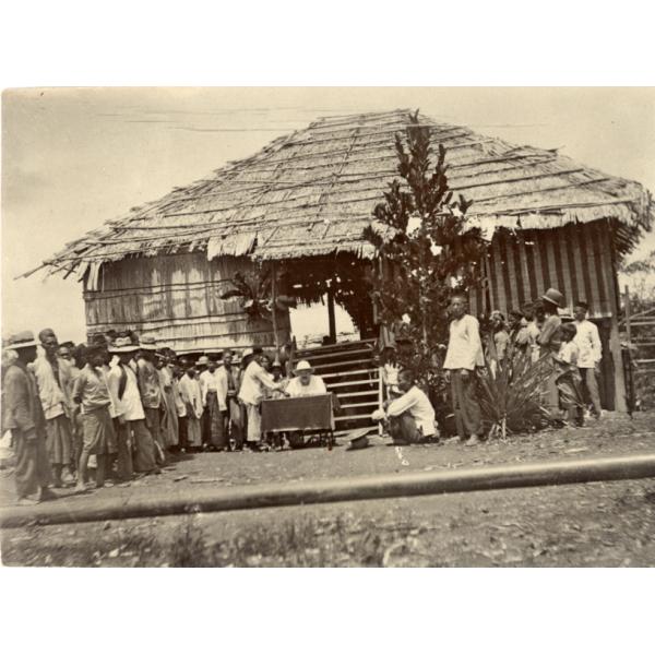 Group of local labourers living up to the left to receive payment from William McRae who is sitting at a desk in front of a hut.