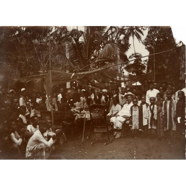 William McRae sitting in a white suit and pith helmet, surrounded by a group of locals under an awning. 