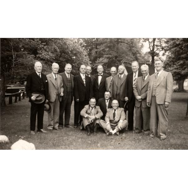 A group of retired International Drillers. Eleven are standing and three are sitting in front on grass. They are all wearing suits. There are trees behind them.