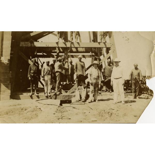A photo of a group of men standing in front of the base of a wooden derrick. There are two International Drillers standing in the front, wearing white outfits and pith helmets.