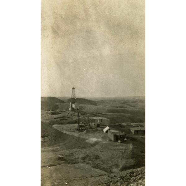 A photo of an oil derrick with four legs, surrounded by rolling hills. There are three small buildings in front of it with pipes running to the derrick.