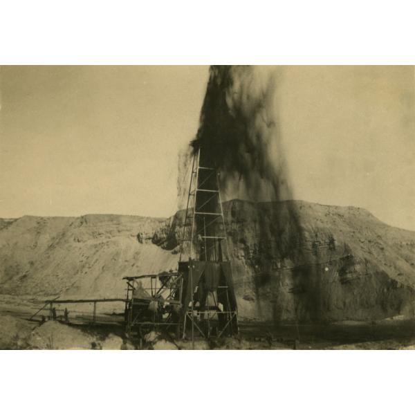 A photo of oil shooting over the top of a derrick with dark curtains around its base. There are dirt hills in the background.