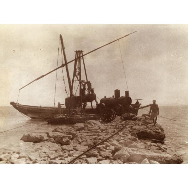 A photo of a boat in the water at the end of a stone pier. There is a crane lifting a steam boiler onto the rocks, and a man walks on the edge of the pier watching the boiler.