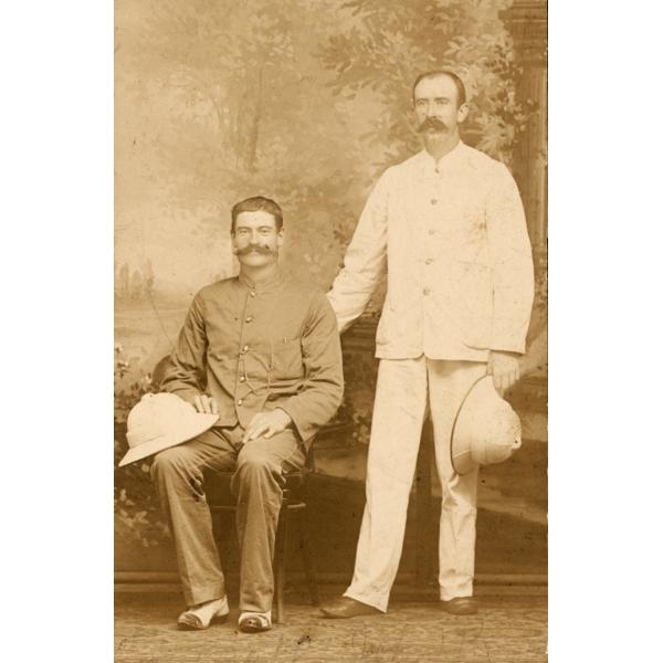A photo of two International Drillers in front of a painted backdrop. On is sitting and one is standing. They are holding white pith helmets and are wearing  button-up shirts.