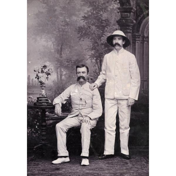 A photo of two men in front of a painted backdrop of trees. They are wearing white suits. There is a table beside the man sitting on the left, with books and flowers on it. The man on the right is wearing a white hat.