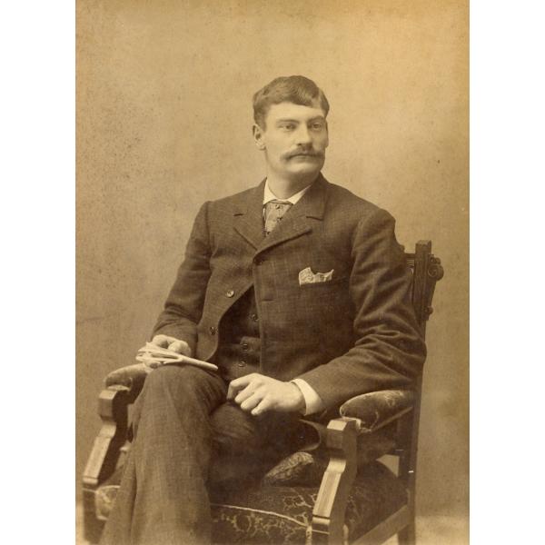 A photo of Thomas Knapp. He is sitting in an arm chair with a plush seat and arm rests. He is holding a folded piece of paper and is wearing a dark suit with the top button of his jacket done up.