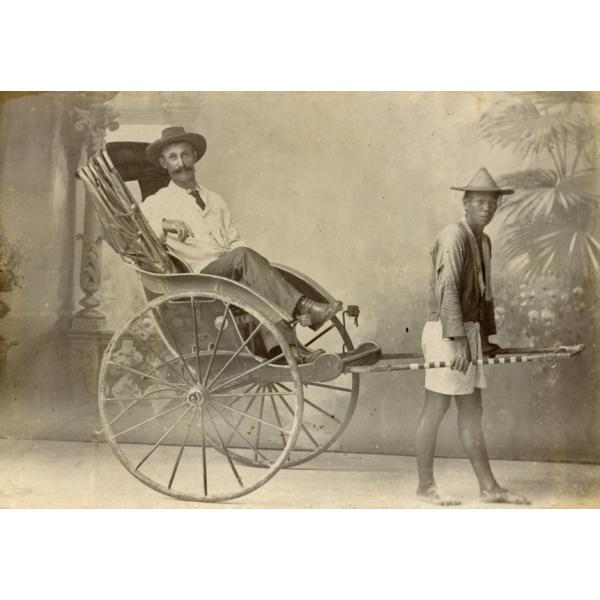 Une photo d'un homme assis dans un pousse-pousse à deux roues avec l'auvent refermé. Il est tiré par un homme de la région qui porte un chapeau de paille. Une toile de fond peinte se trouve derrière eux, avec des piliers et des arbres. 