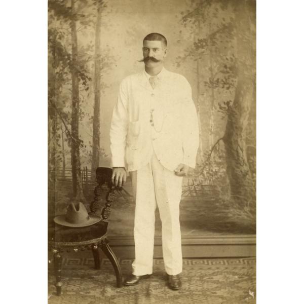 A photo of Thomas Knapp in front of a painted background of trees. He is wearing white and is resting his hand on the back of a chair with a hat on the seat.