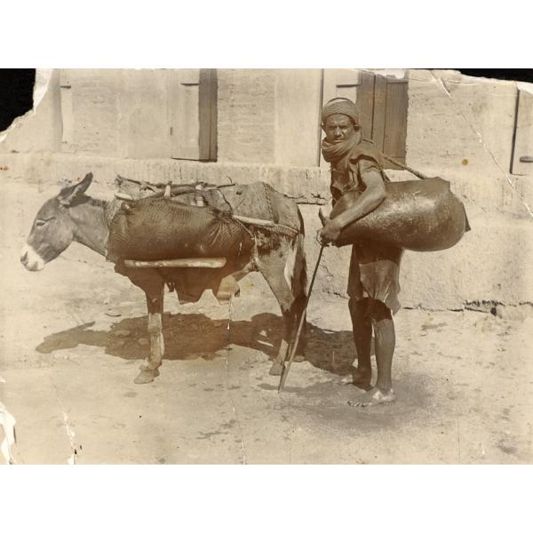 A photo of a man carrying a large sack beside a grey donkey. The man is holding a cane and the donkey has a wooden frame on its back, holding a bundle. They are in front of a stone building.