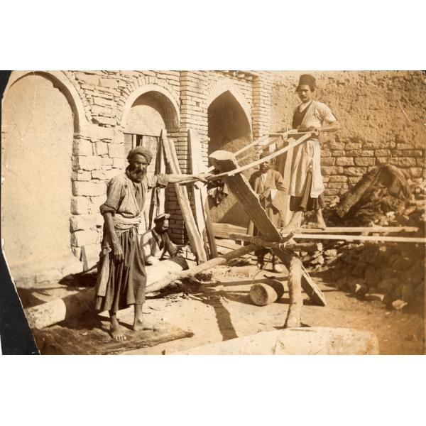 Un groupe de quatre hommes debout devant un mur de pierres avec des arches dans celui-ci. Deux d'entre eux utilisent une scie pour couper un morceau de bois qui est appuyé sur un cadre en bois. 