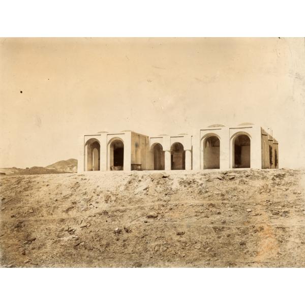 A photo of a concrete bungalow on a hill in the desert. There are two wings coming off of the centre part, each with two open arches. There are three arches in the middle section.