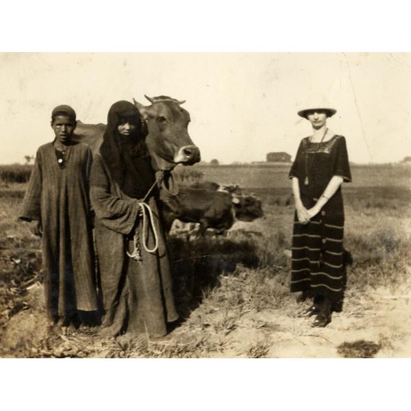 A photo of Annie Dyke standing beside a boy and woman holding a cow. Annie is wearing a dark striped dress, the woman a dark robe and head covering, and the boy a dark robe. There is a field behind them.