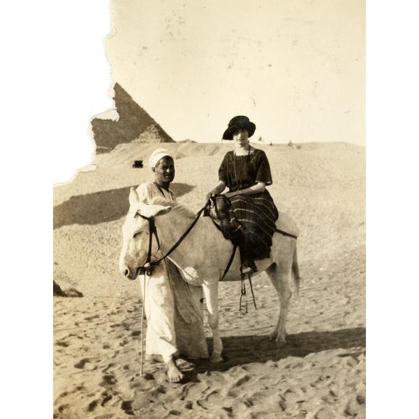 A photo of Annie Dyke in the desert, sitting on a white donkey that is being held by an Egyptian man. She is wearing a dark striped dress and hat, while he is in a white robe. There is a pyramid in the background.