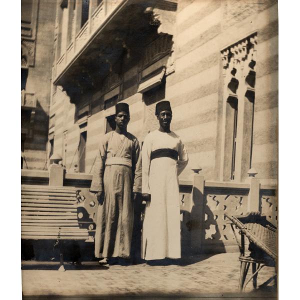 Une photo de deux hommes portant de longues robes avec des chapeaux foncés et des bandes autour de la taille. Ils sont debout à côté d'un banc devant un bâtiment en pierres rayées.