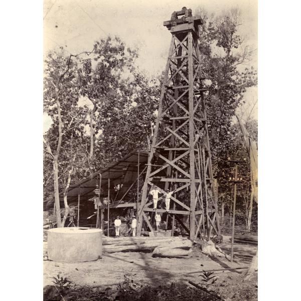 A photo of a group of International Drillers standing at the base and on the first support beam of an oil derrick. They are wearing white. There is a large cylinder and some logs in front of them and trees in the background.