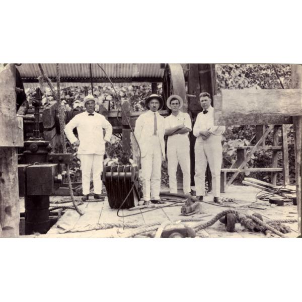 Un groupe de quatre hommes debout à l'intérieur d'une tour de forage pour le pétrole sur un plancher fait de planches de bois. Ils sont vêtus de blanc. Une grande roue se trouve derrière eux et des machines se trouvent à côté d'eux. 