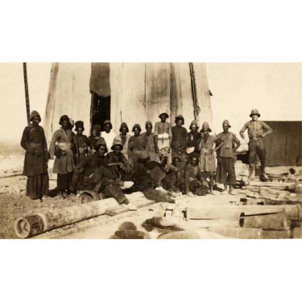 A photo of a crew of labourers with an International Driller who is wearing a pith helmet and standing on the right. They are in front of a derrick covered in cloth. There is a pile of casing in front of them.