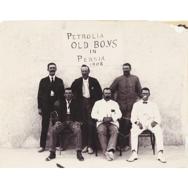Six men in suits. Three are standing and three are sitting. There is a sign on the wall behind them that says, "Petrolia Old Boys in Persia 1908".