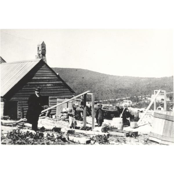 Charles Wallen standing in an oil field in front of a wooden building. There are workers and machinery behind him. There is a hill in the background covered with trees. 