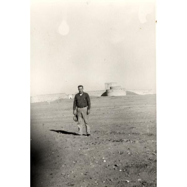 Une photo d'un homme debout dans le désert, tenant son chapeau à ses côtés. Derrière lui, il y a des murs de pierre avec une tour circulaire à l'avant et une tour carrée plus haute derrière.  