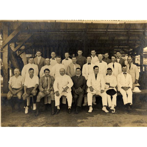 Group photograph of foreign drillers in Borneo all wearing white suits. They are sitting or kneeling on a raised platform underneath a roofed structure. 