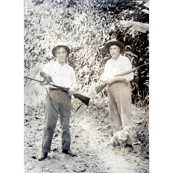 Deux hommes vêtus de chemises blanches, de chapeaux et de pantalons foncés. Ils portent des fusils. Il y a un petit chien au pied de l'homme à droite. Ils sont debout devant un mur de végétation. 