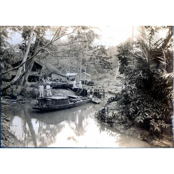 Six men on a boat in a narrow river. There are two men on a dock with a pile of casing beside them on the shore. A group of men stands behind the basing in front of three buildings. There are trees in the background.