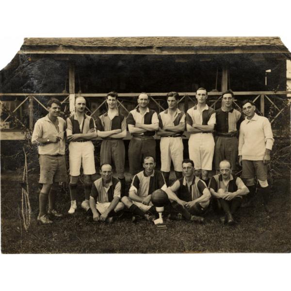 A photo of a group of men wearing matching shirts with black on one side and light on the other. Eight are standing with their arms crossed. Four are sitting cross-legged with a ball on a stand in front of them.