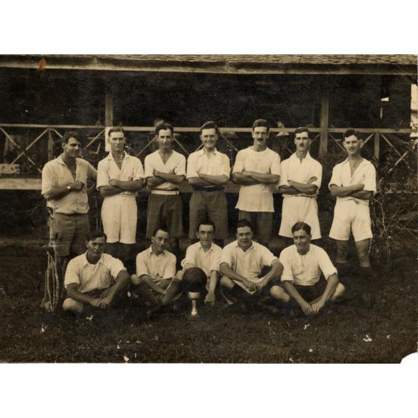 A photo of a group of men wearing white shirts. Seven are standing with their arms crossed. Five are sitting cross-legged with a ball on a stand in front of them.