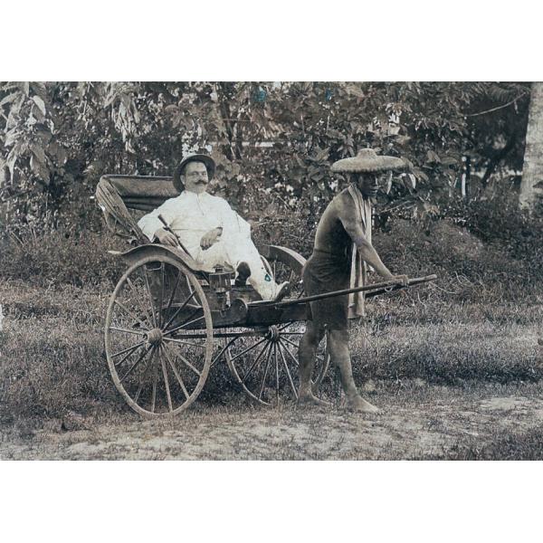 Un foreur international portant un chapeau et un costume blanc dans un pousse-pousse à deux roues tiré par un homme de la région. L'homme porte un grand chapeau de paille et un morceau de tissu autour de ses épaules. La voilure du pousse-pousse est fermée. Ils sont devant des arbres. 